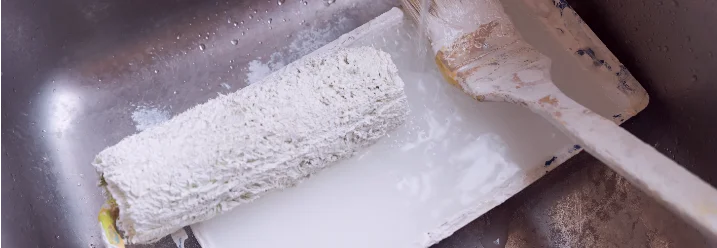 Eine Farbrolle und ein Pinsel mit weißer Farbe liegen in einem Waschbecken.